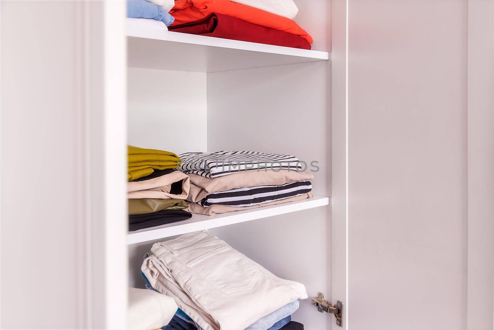 close-up, open white closet with shelves on which lie neatly folded sweaters and shirts. The concept of easy ironing, minimalism, organization of things and space. Optimizing wardrobe items