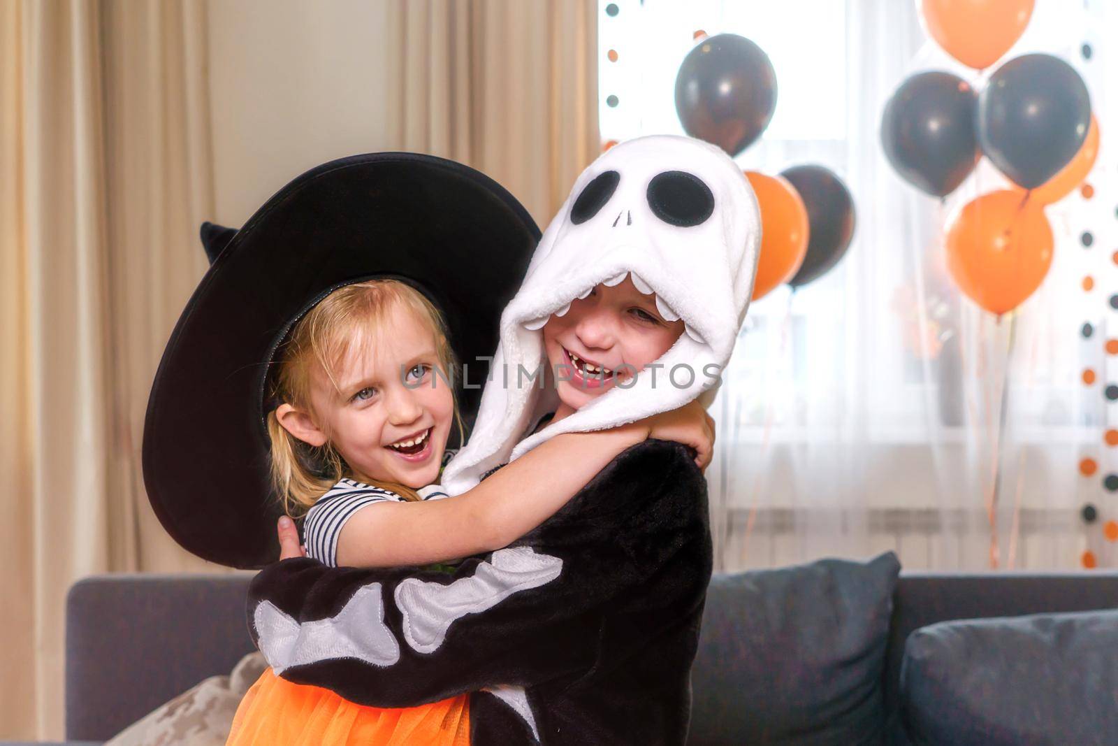 Close-up portrait of boy and girl dressed as a witch and a skeleton. Children hugging and laughing happily, Halloween house party. Against the background of orange decorations and balloons, copy paste