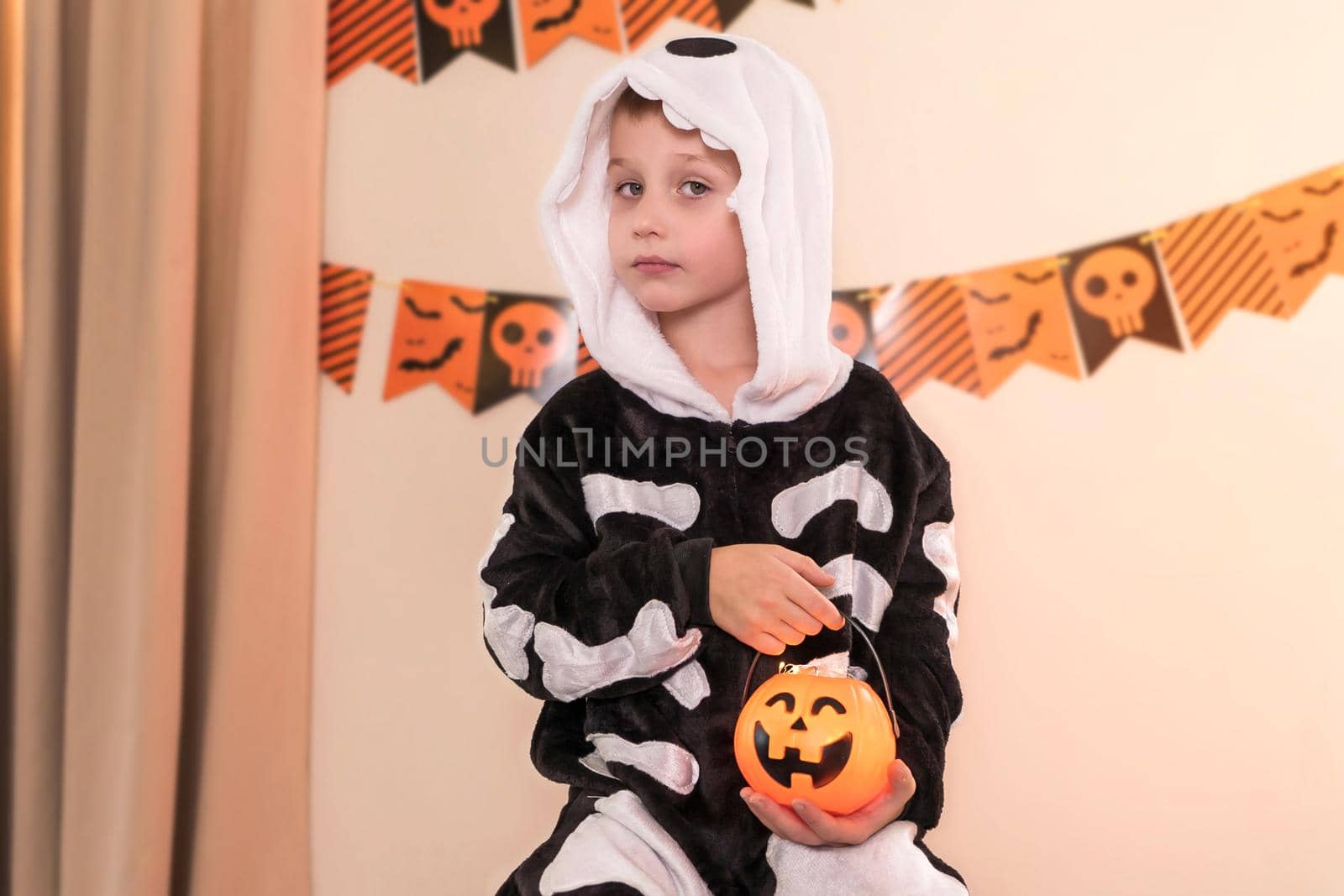 Close-up portrait of a sad boy 5 years old, dressed as a skeleton. Halloween party background. In the hands of a child is a pumpkin lantern with a scary face. The concept of a lonely, sad child