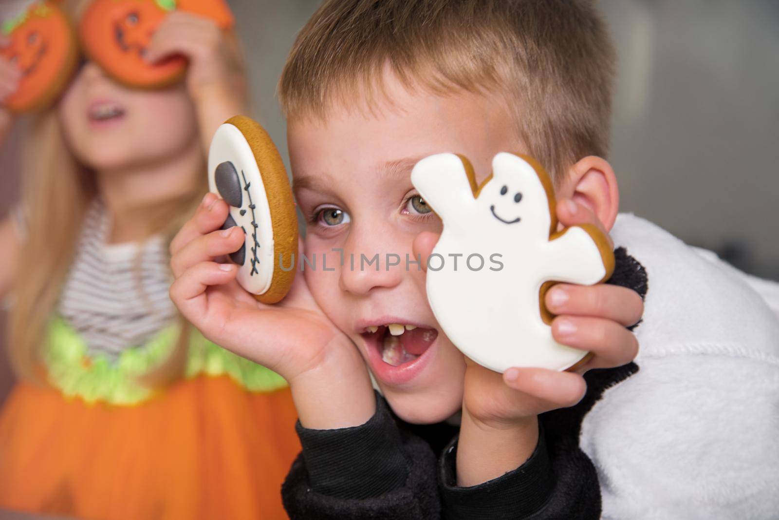 Close-up portrait of children with homemade unusual gingerbread cookies in their hands at the Halloween party. A boy in a skeleton costume, a girl in a pumpkin costume, holding hand-drawn ghost pastry