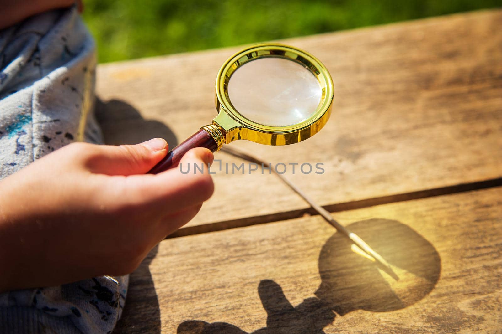 Close-up of a little boy's hand with a magnifying glass on a sunny summer day. The child is trying to light a fire on a wooden bench. The concept of children's experiences, education, curiosity