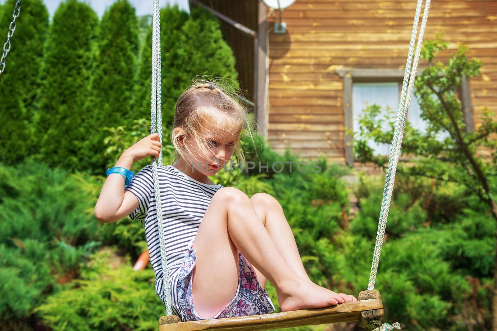 A little girl of European appearance, blonde in a striped T-shirt, sits on a swing. The child is sad, loneliness, no friends. International Children's Day. The concept of child problems. June 1st
