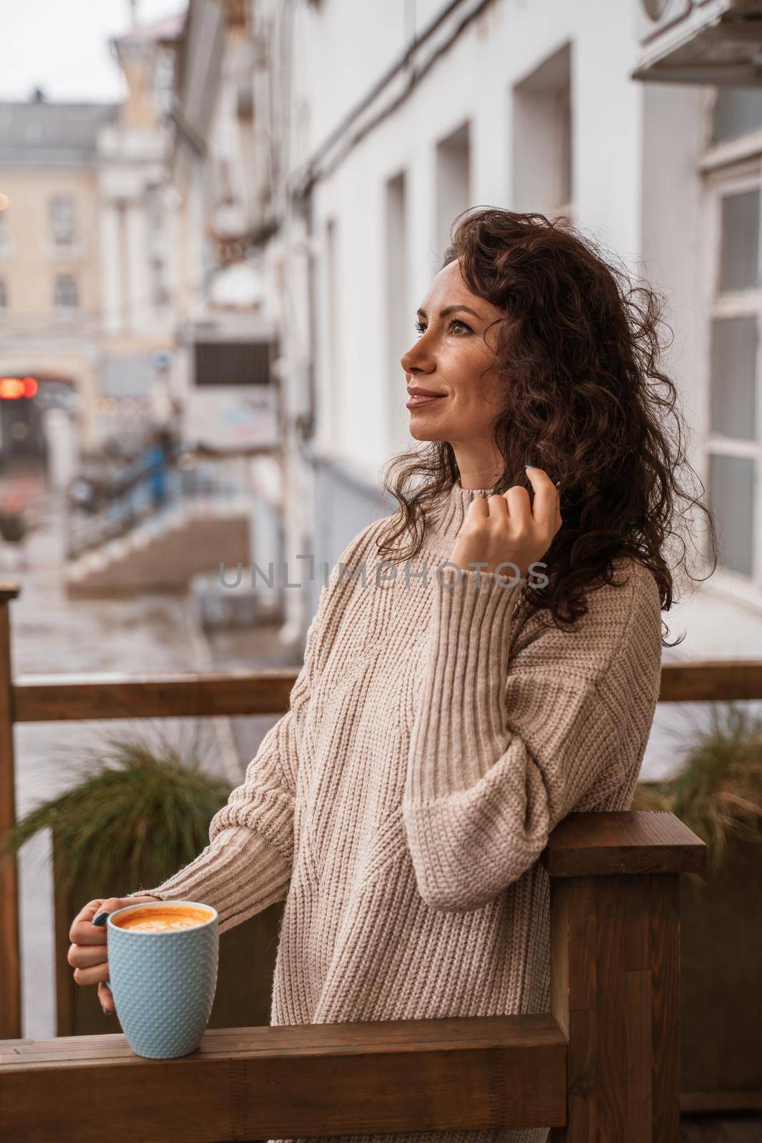 A middle-aged woman in a beige sweater with a blue mug in her hands is in a street cafe on the veranda by Matiunina