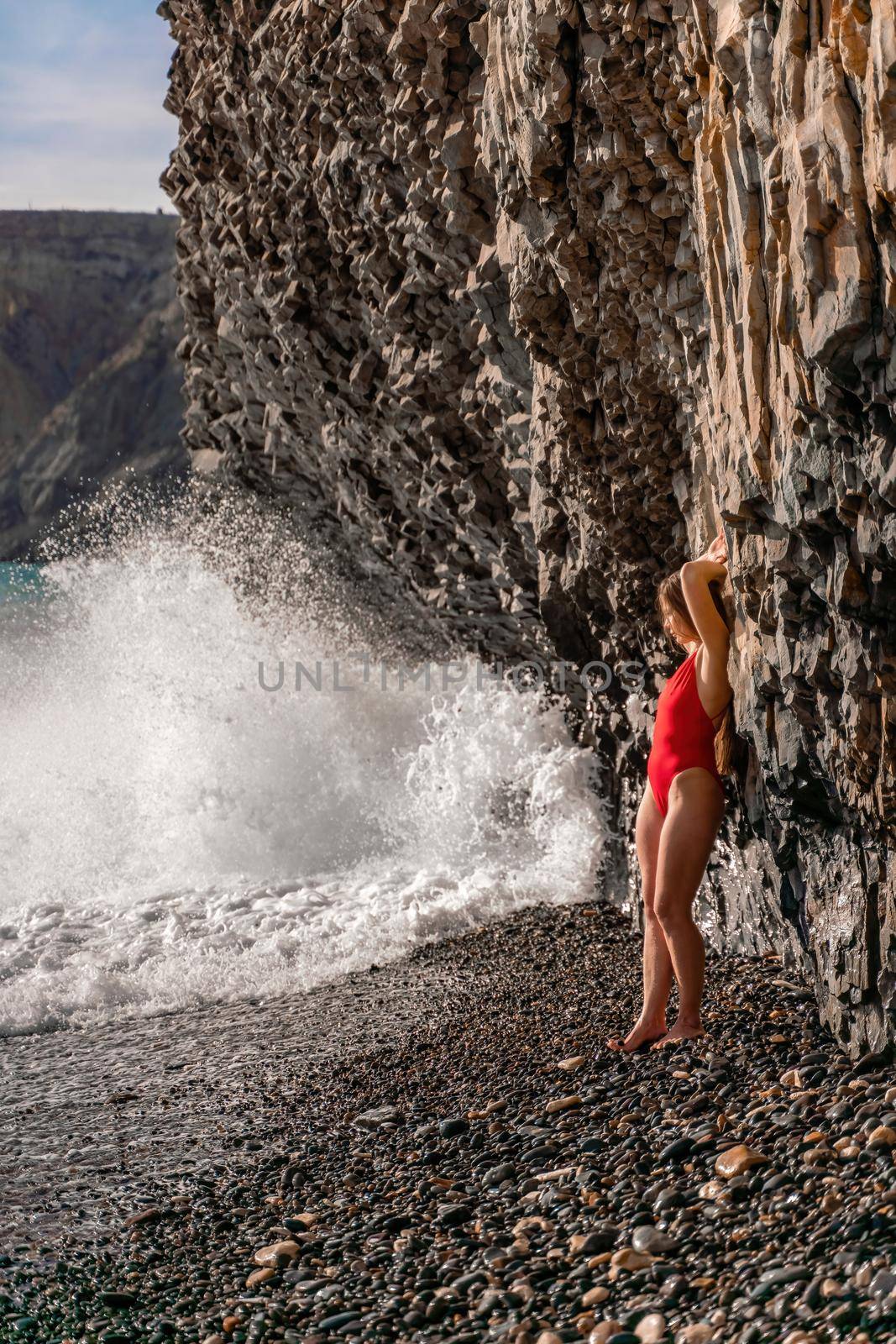 A beauty in a red swimsuit with long legs poses on a fantastic beach with huge waves against the background of mountains.