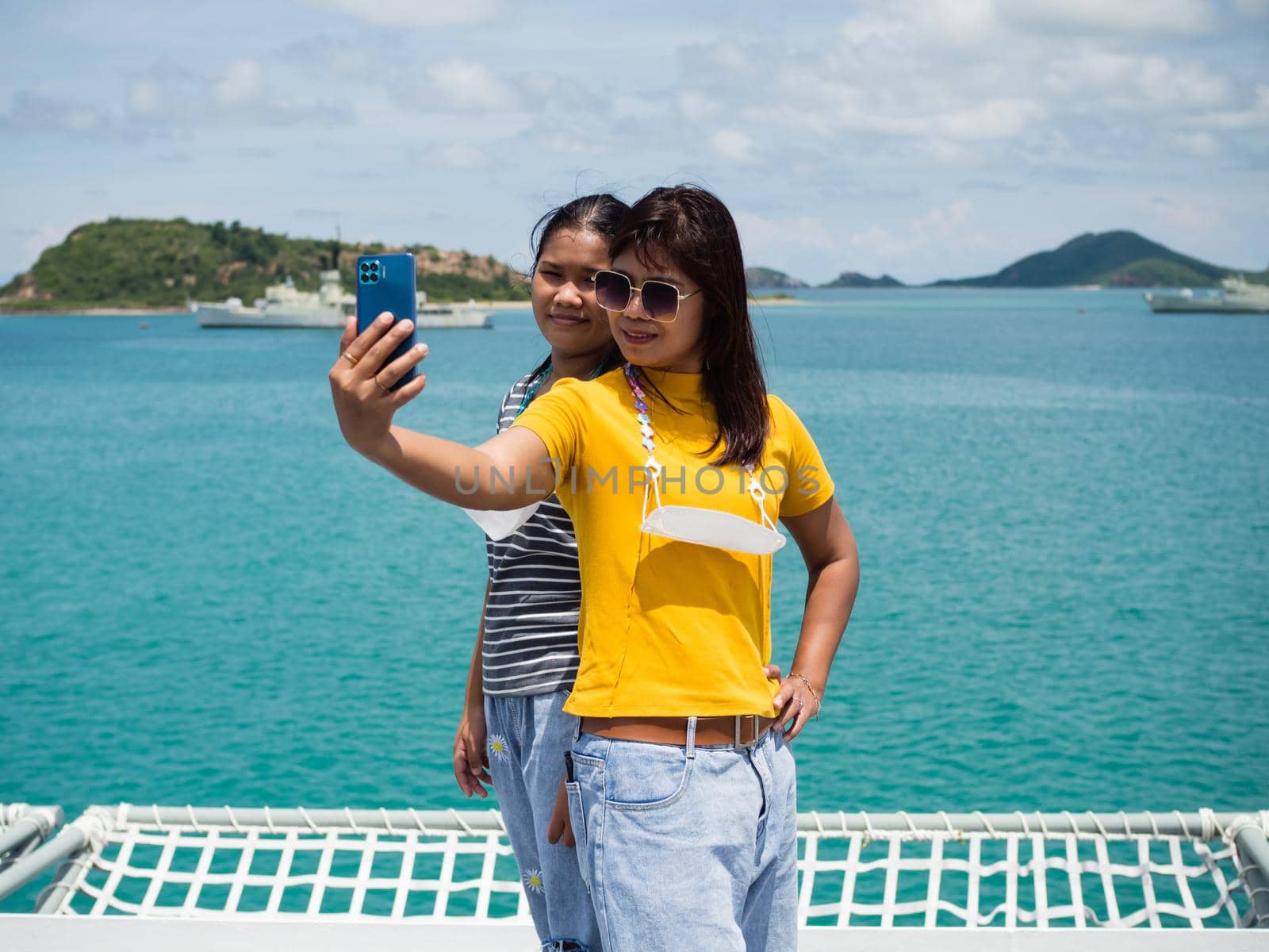 A woman in a yellow shirt is holding a phone to take a selfie with a girl in a gray shirt. with a background of blue water and small mountains. It is tourism after the corona virus outbreak.