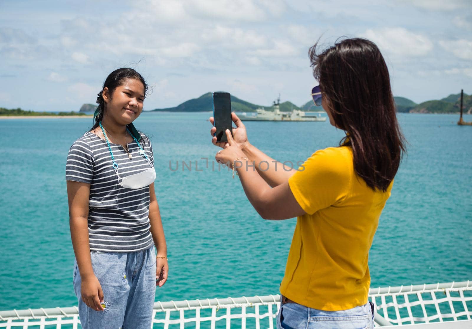 A woman in a yellow shirt is holding a phone to take pictures of a girl in a gray shirt. With a background of blue water and small mountains. It is tourism after the corona virus outbreak.