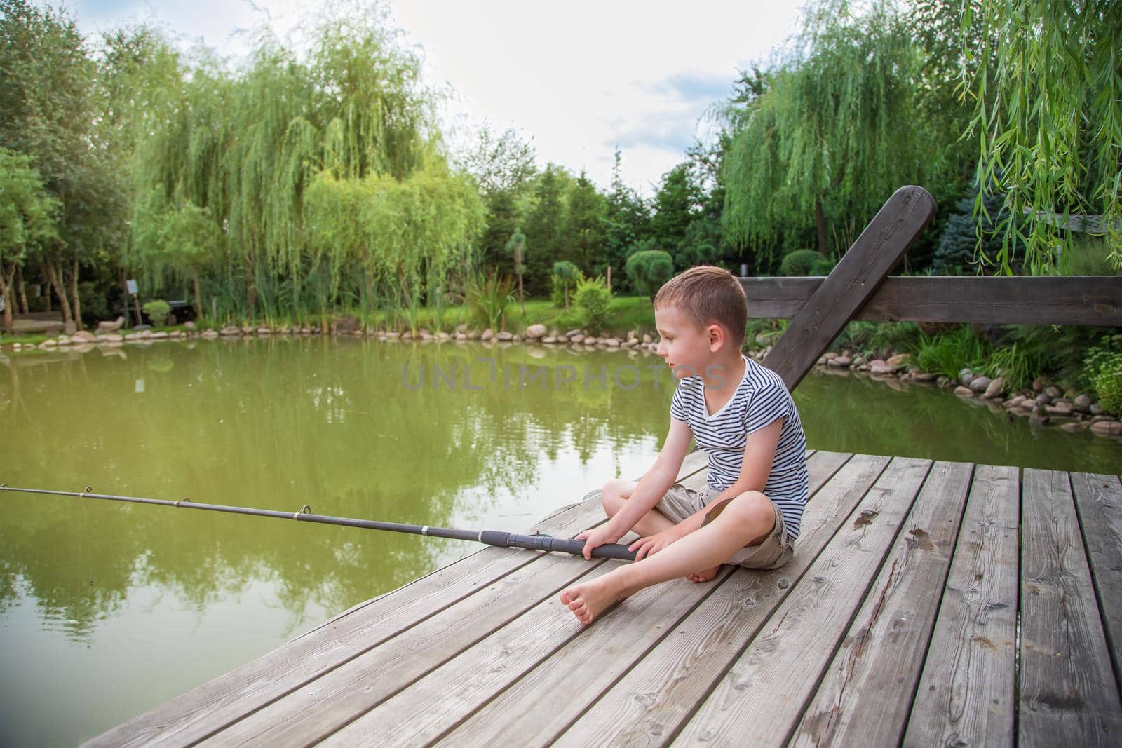 on the shore near the river on sunny day, sad boy sits and catches fish. The child is dressed in a striped T-shirt and shorts, sits on a wooden pier and looks into the distance, holding a fishing rod