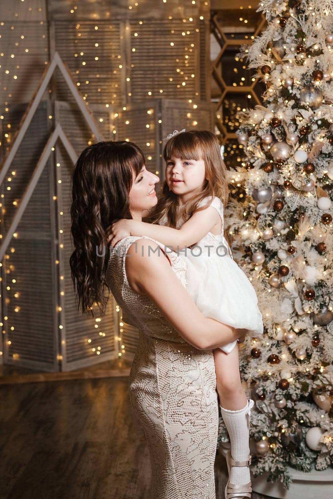 A little girl with her mother in light festive dresses next to the Christmas tree. The theme of New Year's holidays and festive interior with garlands and light bulbs.