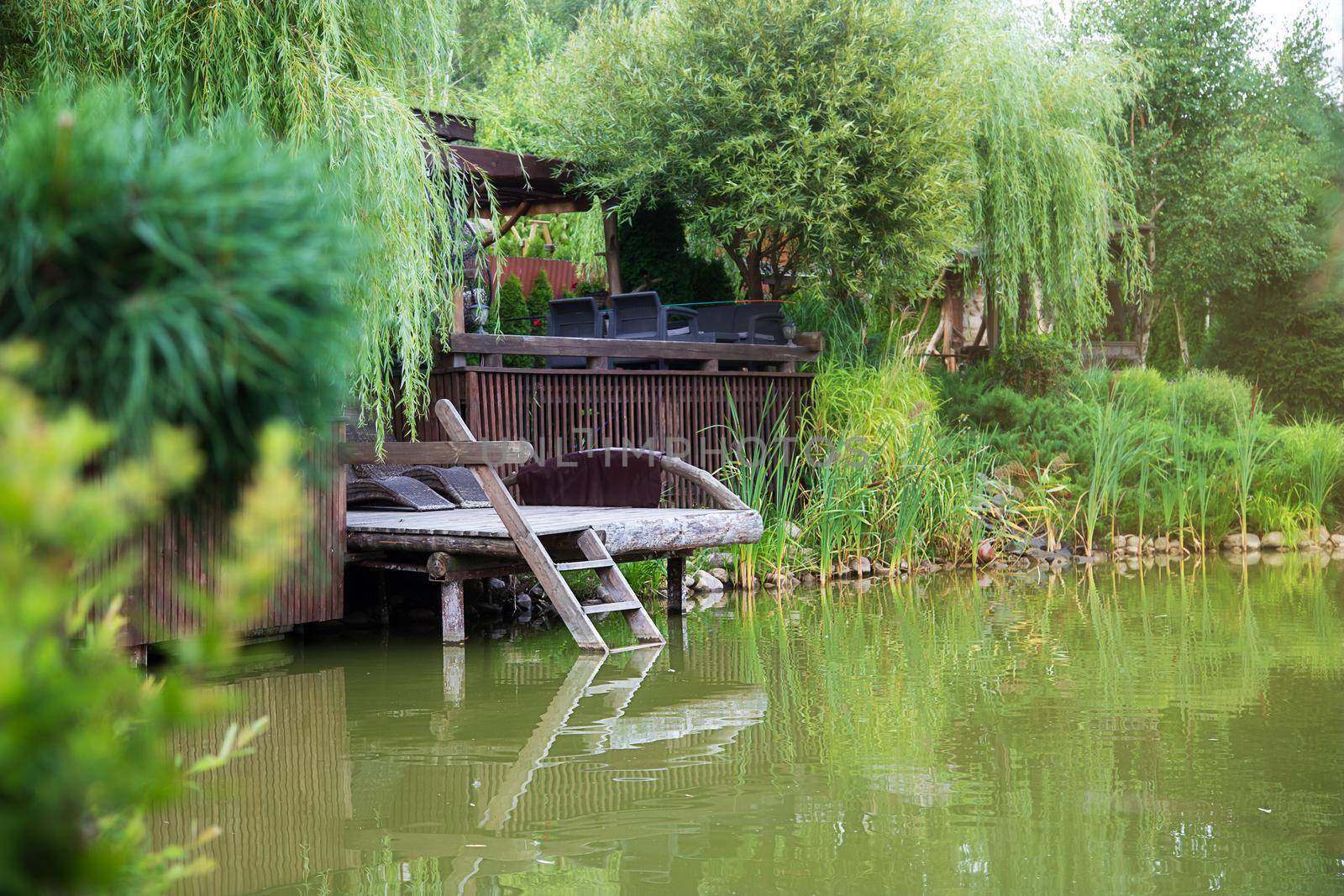 Beautiful summer country landscape. Rustic view of a small lake surrounded by many green trees. Wooden empty jetty with a small ladder. The concept of a calm quiet peaceful life, relaxation, meditation