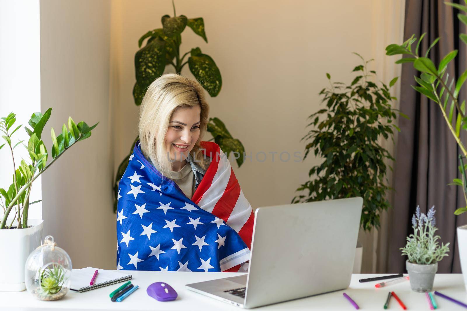 Happy woman employee sitting wrapped in USA flag, shouting for joy in office workplace, celebrating labor day or US Independence day. Indoor studio studio shot isolated on yellow background.