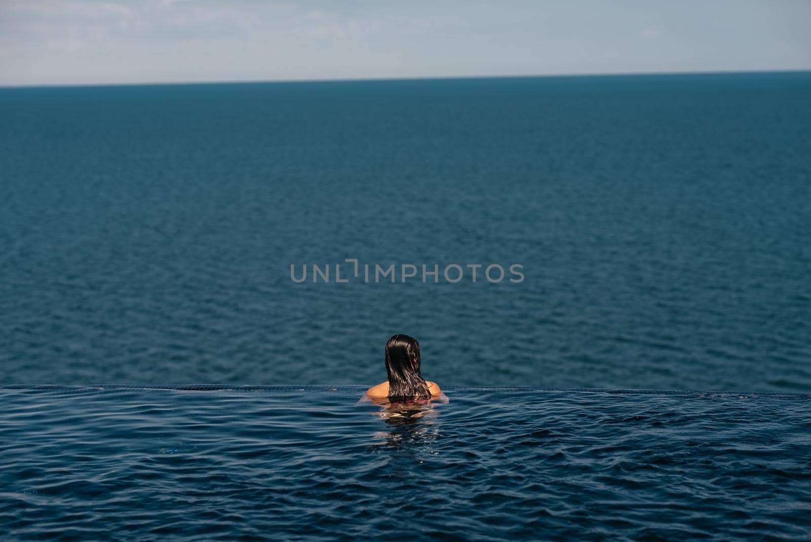 Happy woman in swimsuit swimming in infinity pool at luxury hotel against seafront. Relaxing, summer, travel, holiday, vacation and weekend concept
