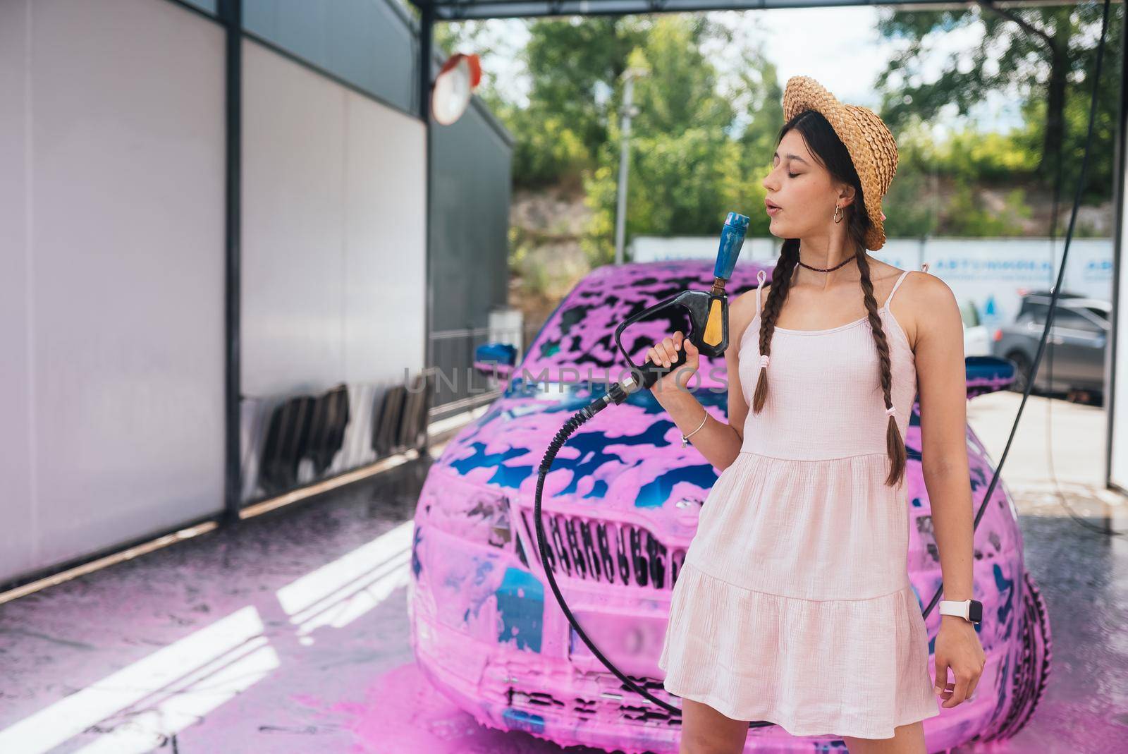 Young woman high pressure hose stands in front of a car covered in pink foam