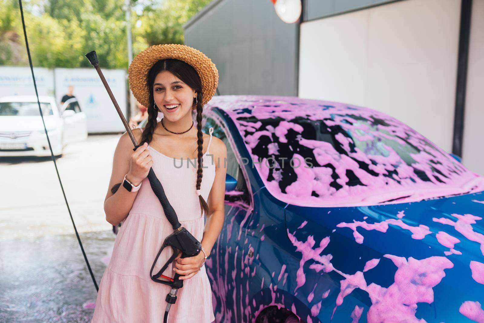 Young woman with high pressure hose stands by car covered in pink foam