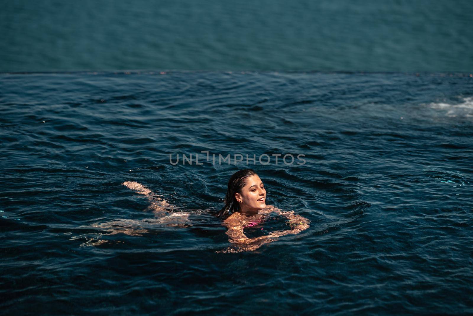 Happy woman in swimsuit swimming in infinity pool at luxury hotel against seafront. Relaxing, summer, travel, holiday, vacation and weekend concept