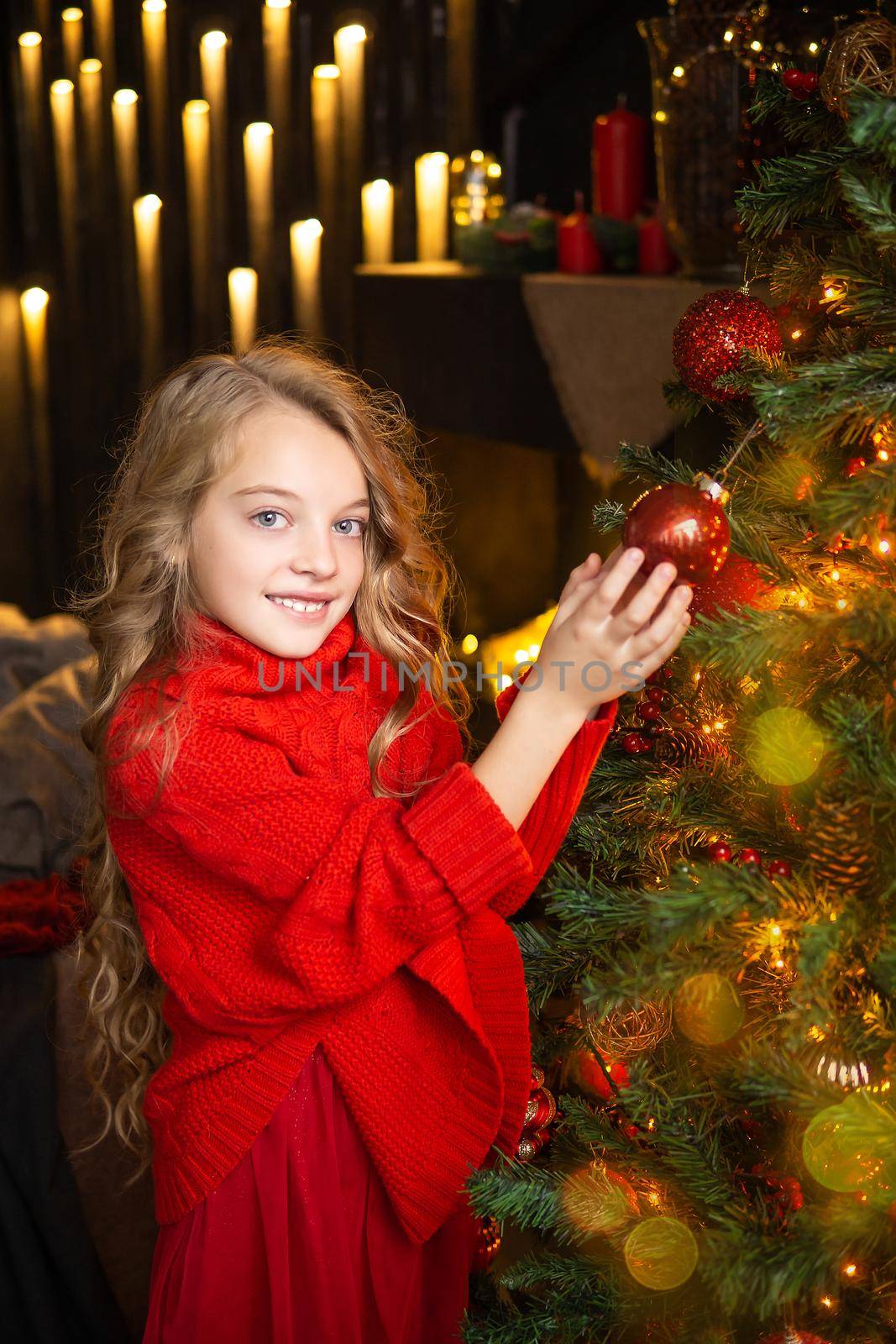 A blonde little girl in a festive outfit next to a Christmas tree decorated with garlands, glass balls and Christmas toys. The concept of winter holidays is Christmas and New Year holidays. Magical festive atmosphere.