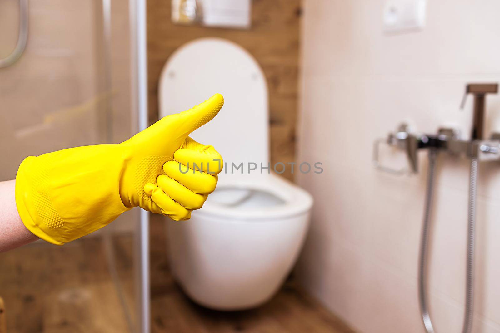 Against the background of a toilet bowl and a bathroom in a modern style, close-up of a hand in a yellow protective glove shows the sign excellent. The concept of good cleaning, indoor hygiene.