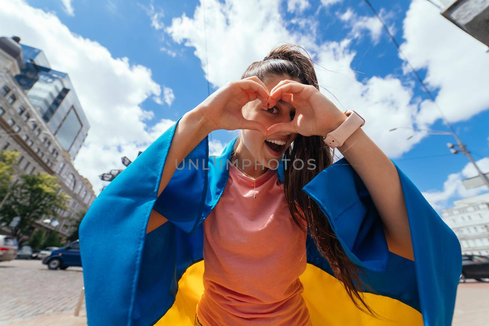 Young woman with Ukrainian flag on shoulders shows heart with hands