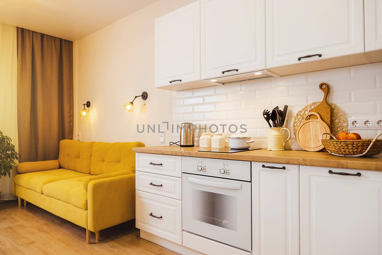 small studio apartment with yellow sofa, white kitchen. Cozy light on by Ramanouskaya