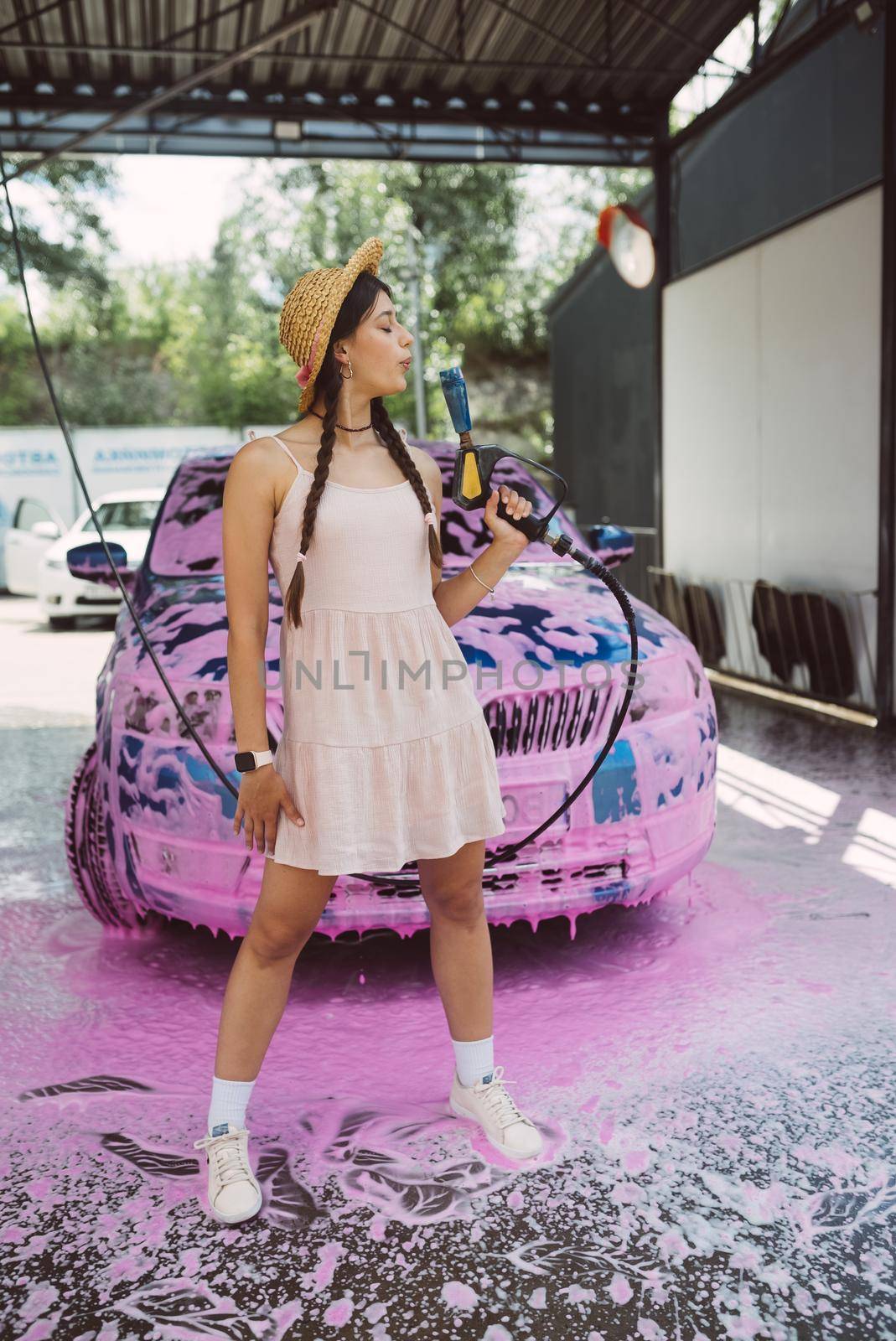 Young woman high pressure hose stands in front of a car covered in pink foam