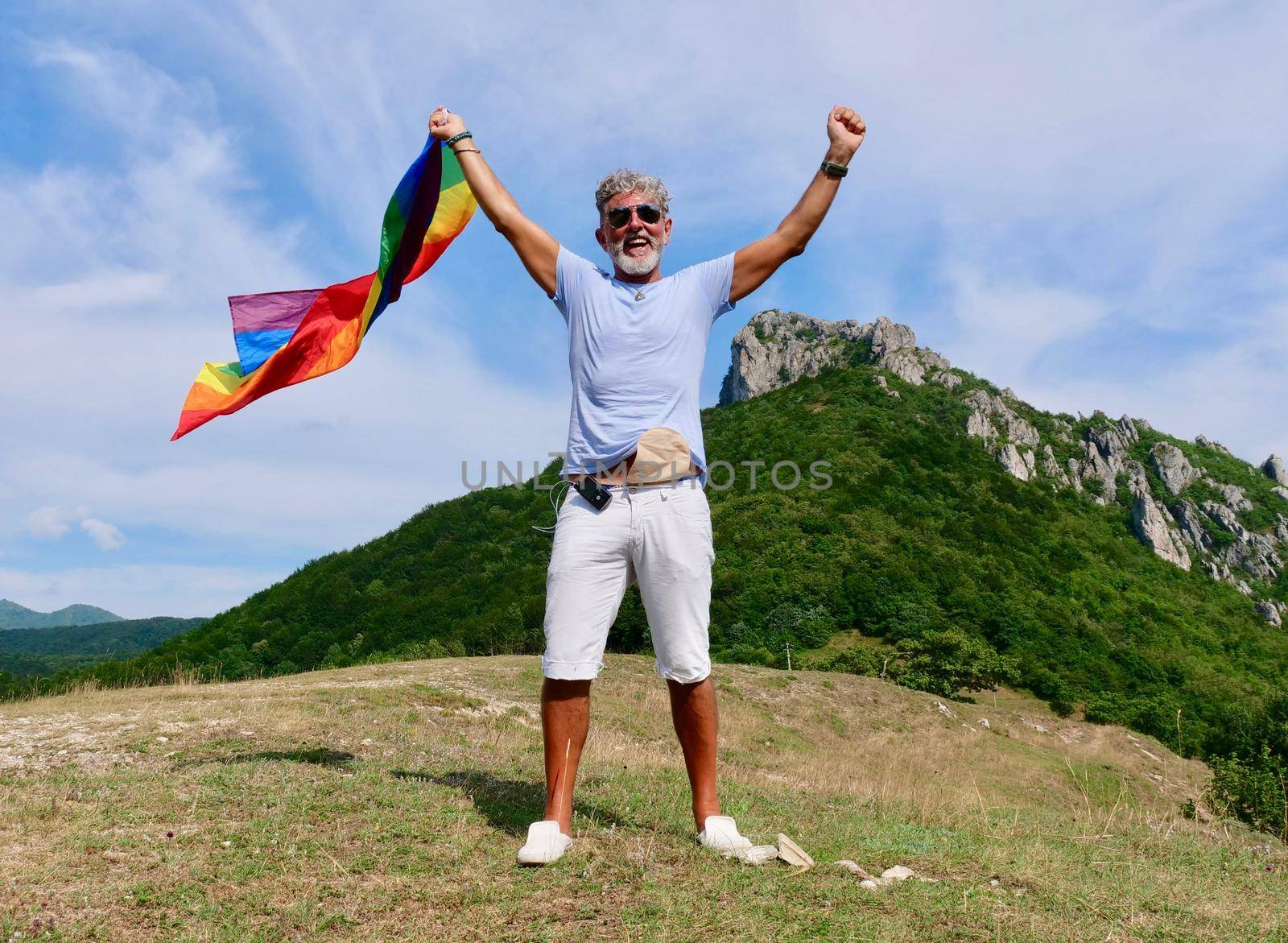 Gray-haired senior elderly man diabetic bisexuality with a beard and sunglasses holding a rainbow LGBTQIA flag on nature. Celebrates Pride Month, Rainbow Flag Day, gay parade