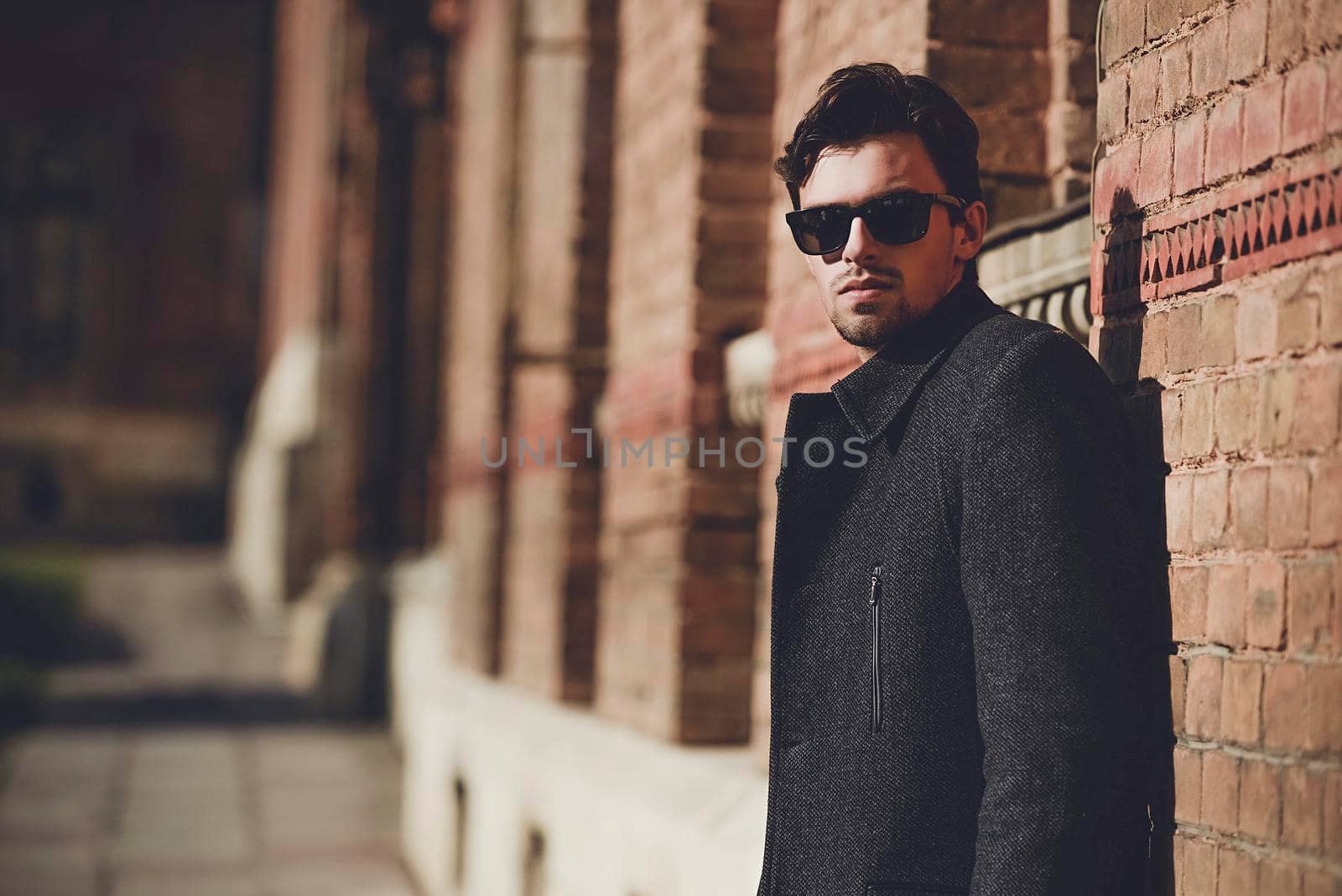 Portrait of fashionable well dressed man with beard posing outdoors looking away, confident and focused mature man in coat standing on a brick wall background, elegant fashion model.
