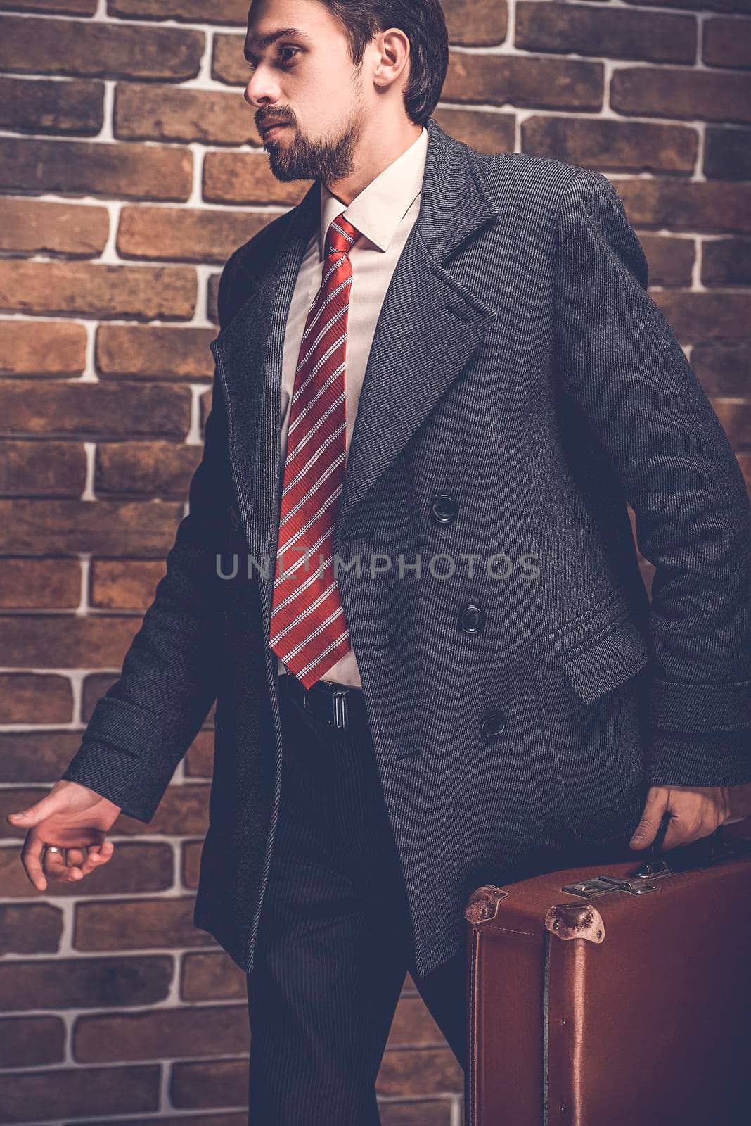 Portrait of fashionable well dressed man with beard posing outdoors looking away, confident and focused mature man in coat standing with suitcase on a brick wall background, elegant fashion model.