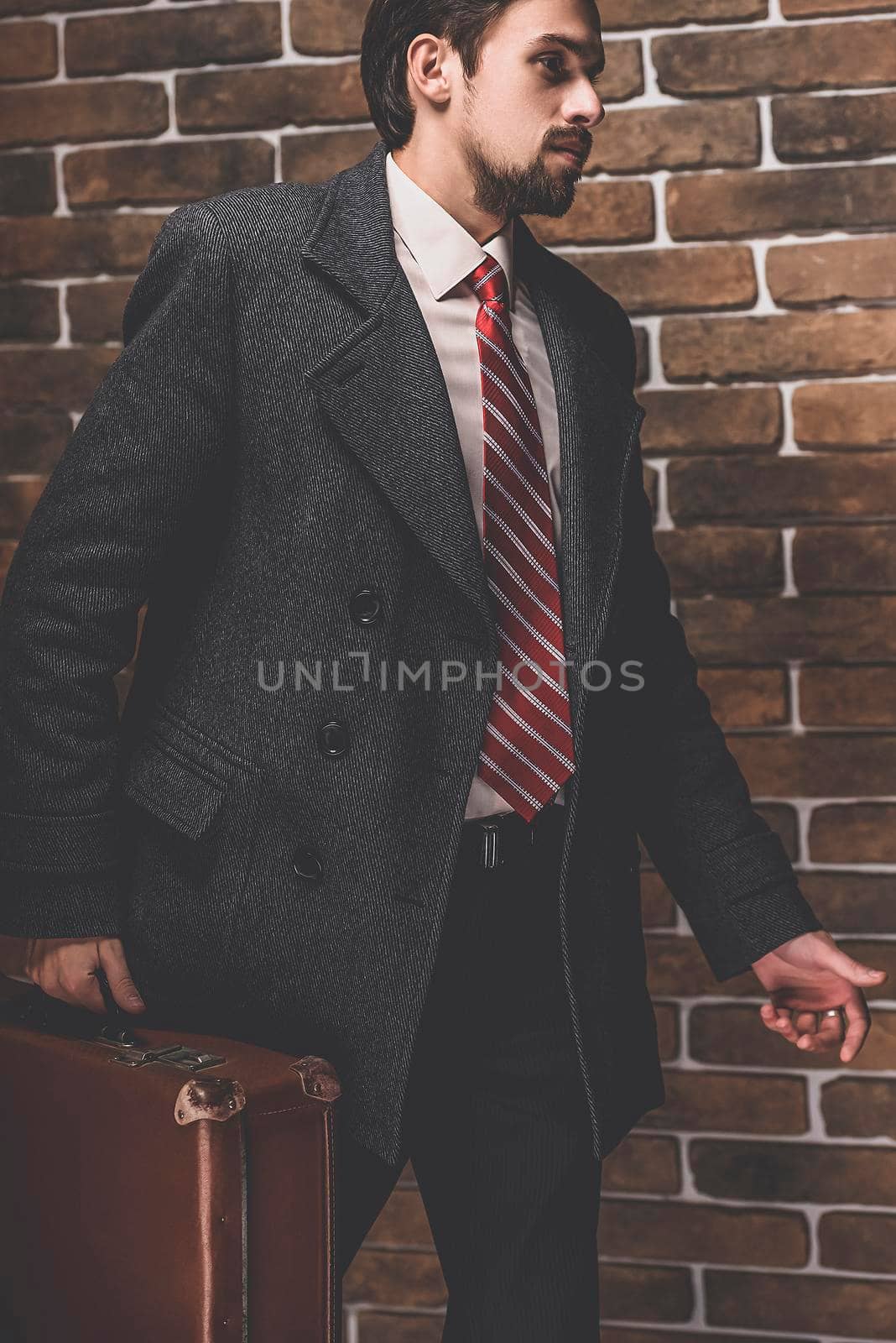 Portrait of fashionable well dressed man with beard posing outdoors looking away, confident and focused mature man in coat standing with suitcase on a brick wall background, elegant fashion model.