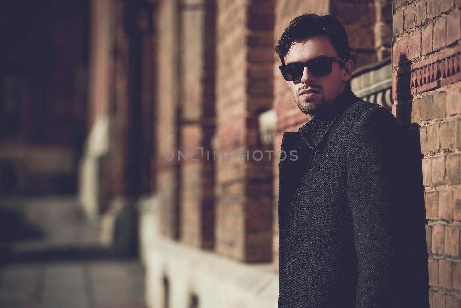 Portrait of fashionable well dressed man with beard posing outdoors looking away, confident and focused mature man in coat standing on a brick wall background, elegant fashion model.