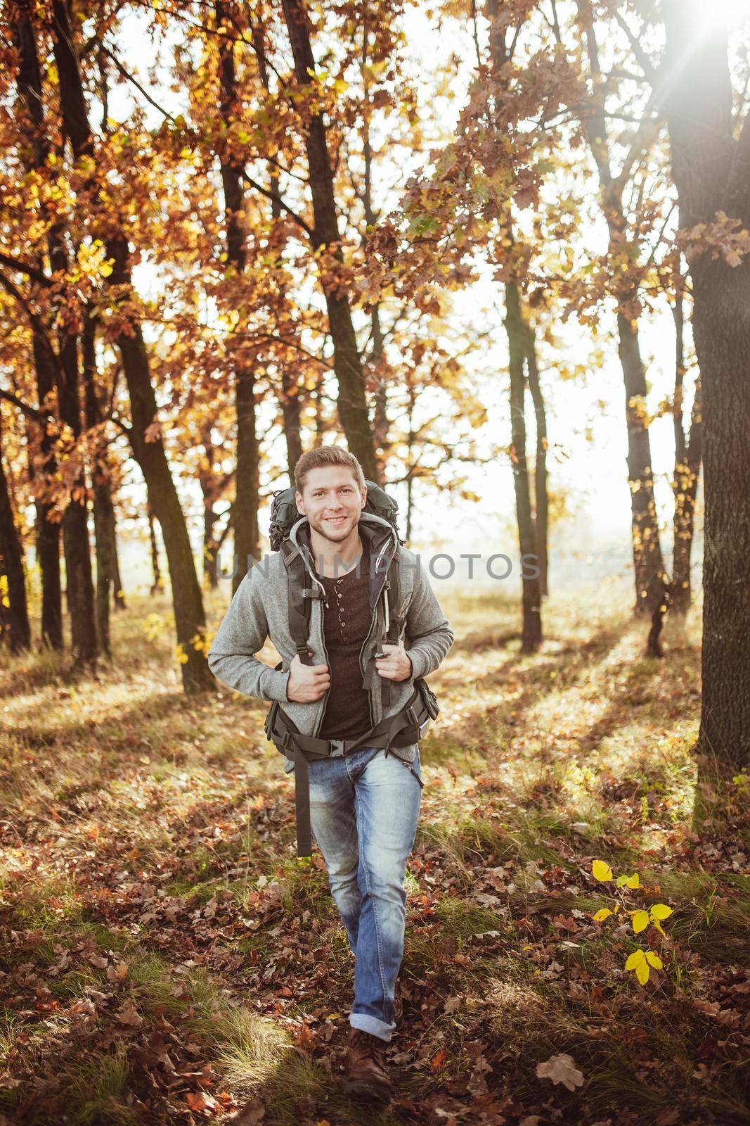 Young man with a backpack travels through the autumn forest on a sunny warm day. Smiling Caucasian tourist walks in nature. Hiking concept.