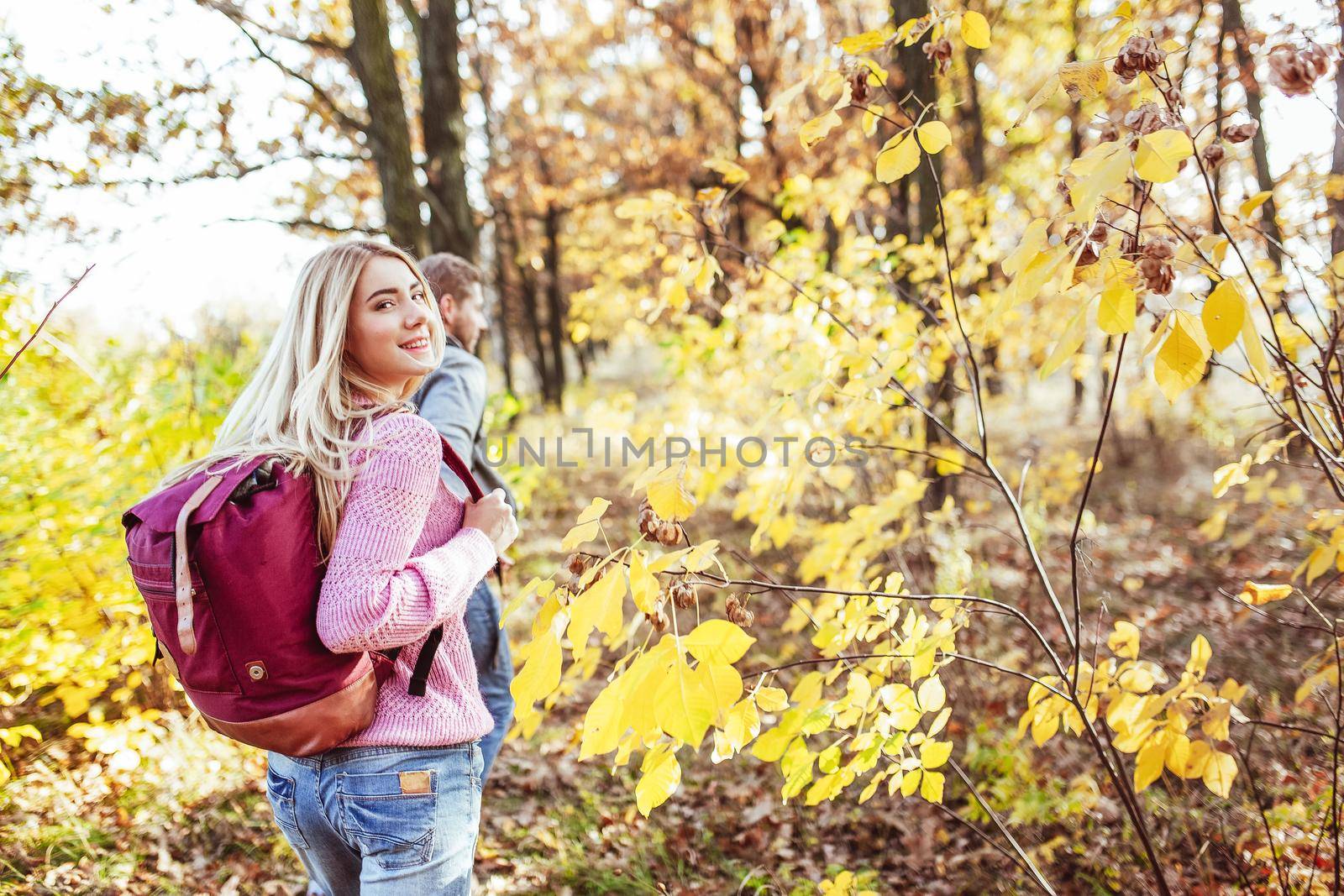 Happy travelers go to the autumn forest holding hands to meet new adventures. Focus on the charming blonde in the foreground who turned around looking at the camera. Togetherness with nature concept.