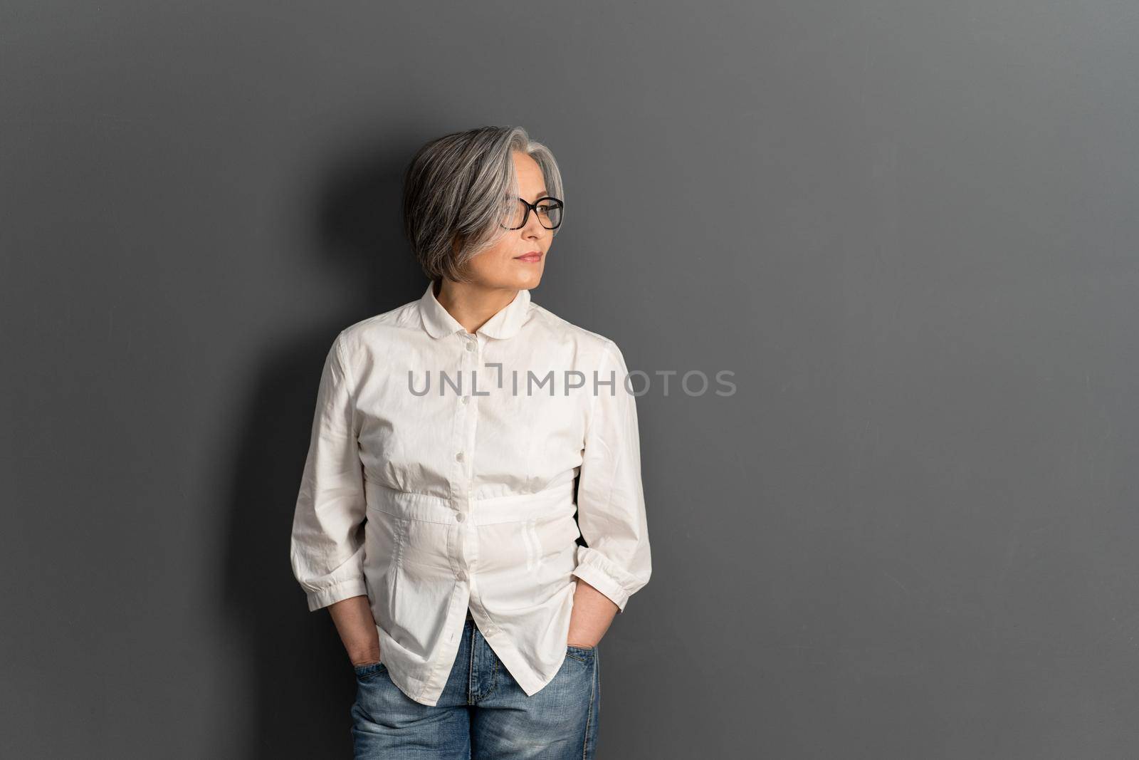 Beautiful mature lady in glasses stands hands in pockets looking at side. Elegant Gray-haired woman in whiite shirt on gray background. Copy space for text at right side by LipikStockMedia