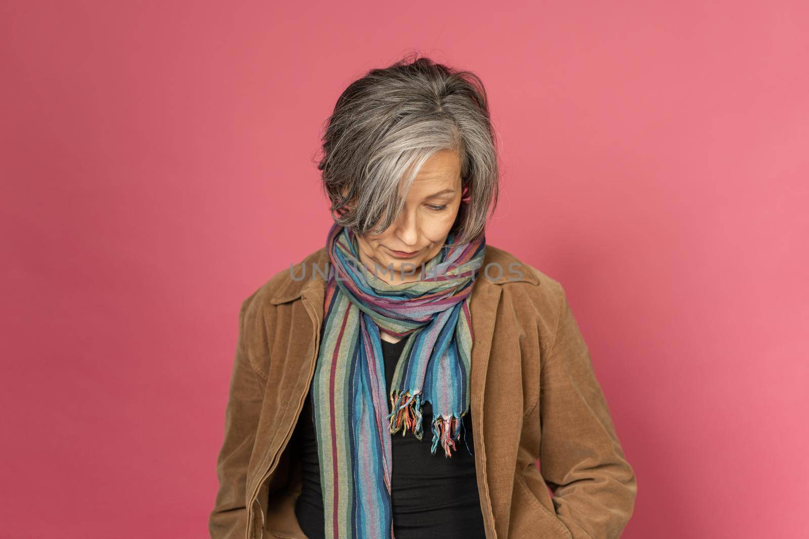 Pensive grey haired woman looks down posing in studio on pink background. Lonely concept.