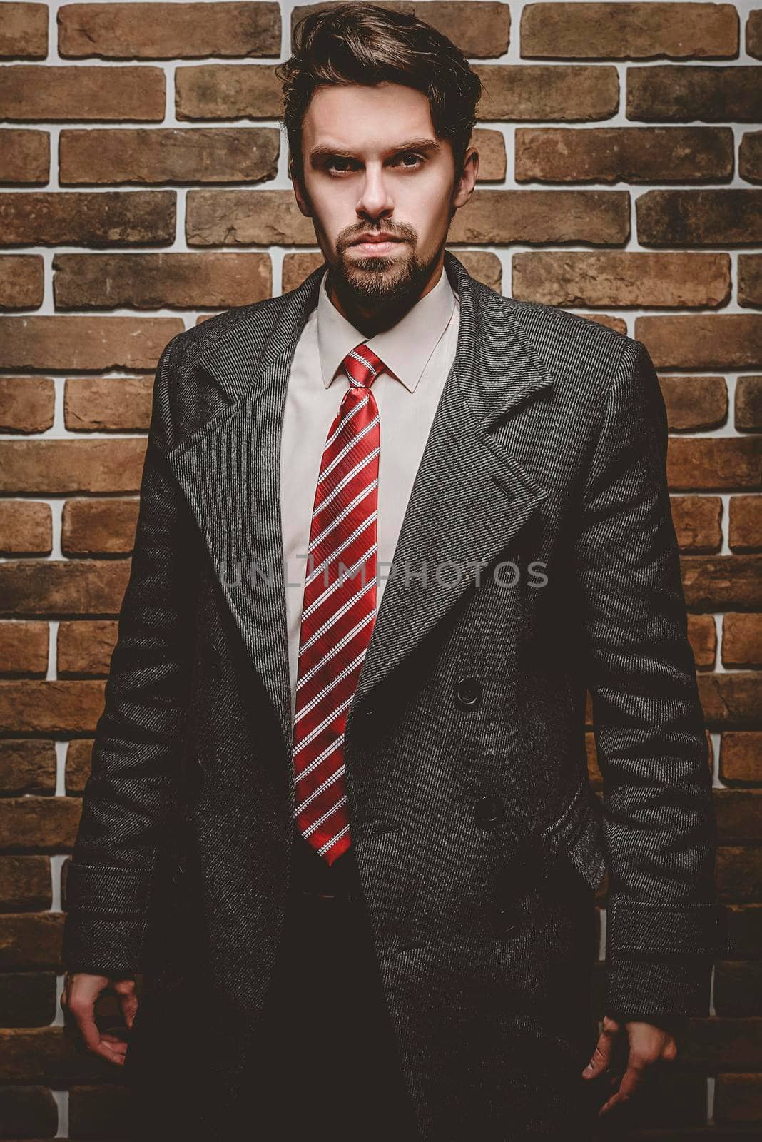 Portrait of fashionable well dressed man with beard posing outdoors looking away, confident and focused mature man in coat standing on a brick wall background, elegant fashion model.