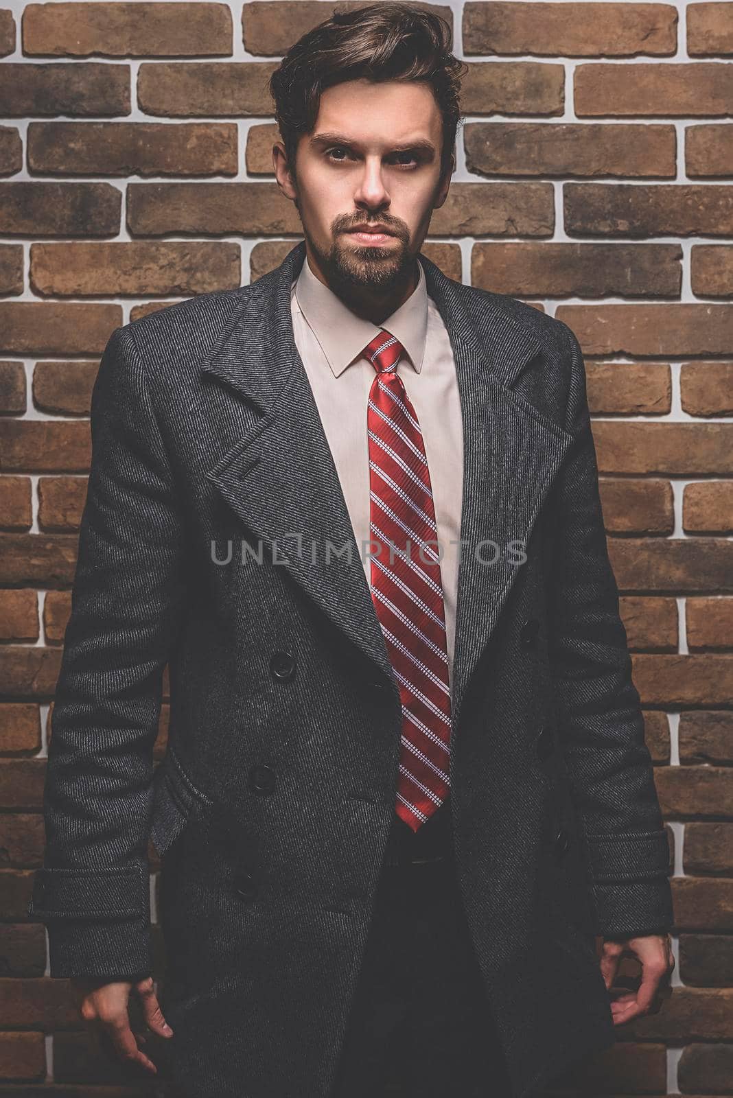 Portrait of fashionable well dressed man with beard posing outdoors looking away, confident and focused mature man in coat standing on a brick wall background, elegant fashion model.