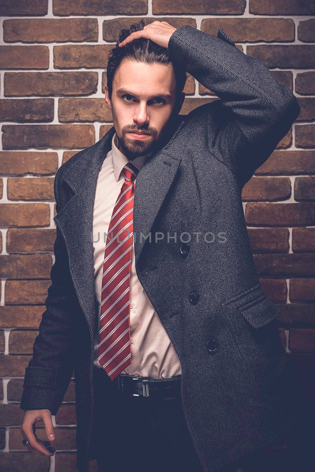 Portrait of fashionable well dressed man with beard posing outdoors looking away, confident and focused mature man in coat standing on a brick wall background, elegant fashion model.