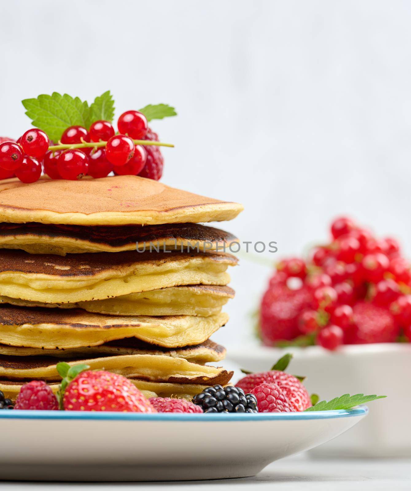 Stack of baked pancakes with fruits in a round plate on a white table