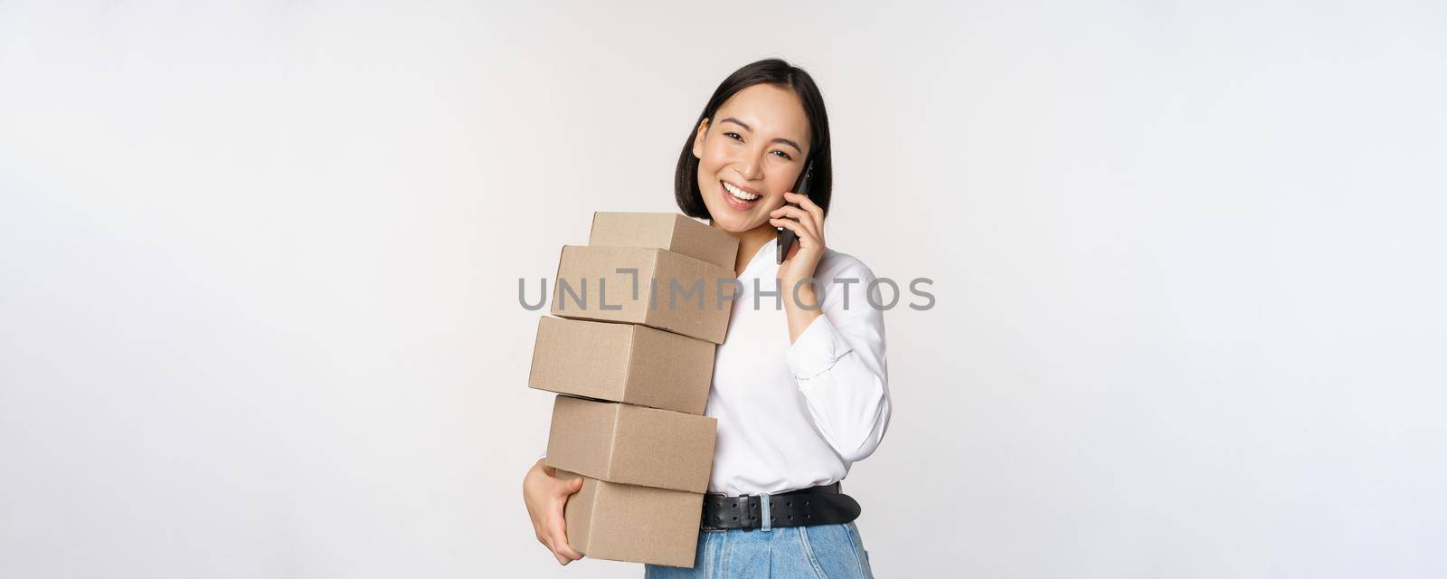 Image of young asian businesswoman answer phone call while carrying boxes for delivery, posing against white background by Benzoix