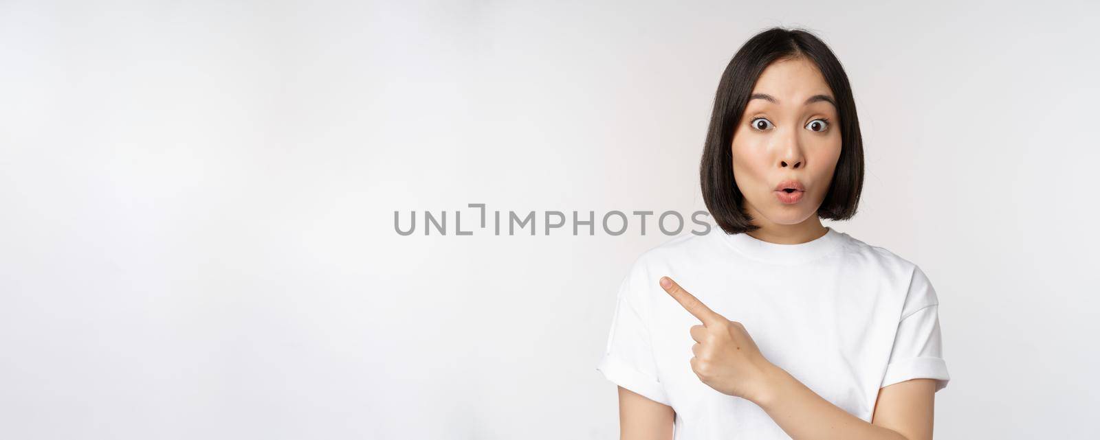 Close up of young japanese woman smiling, pointing finger left at copy space, showing announcement or advertisment banner, standing over white background by Benzoix