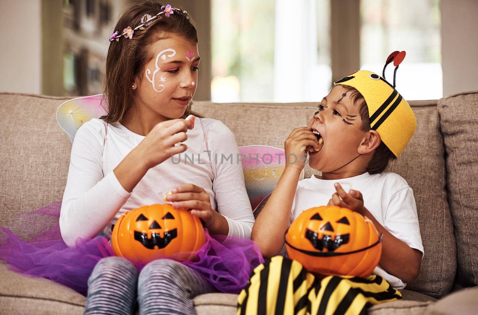 Dont eat it all at once. a young brother and sister eating their Halloween candy while sitting on their sofa at home. by YuriArcurs