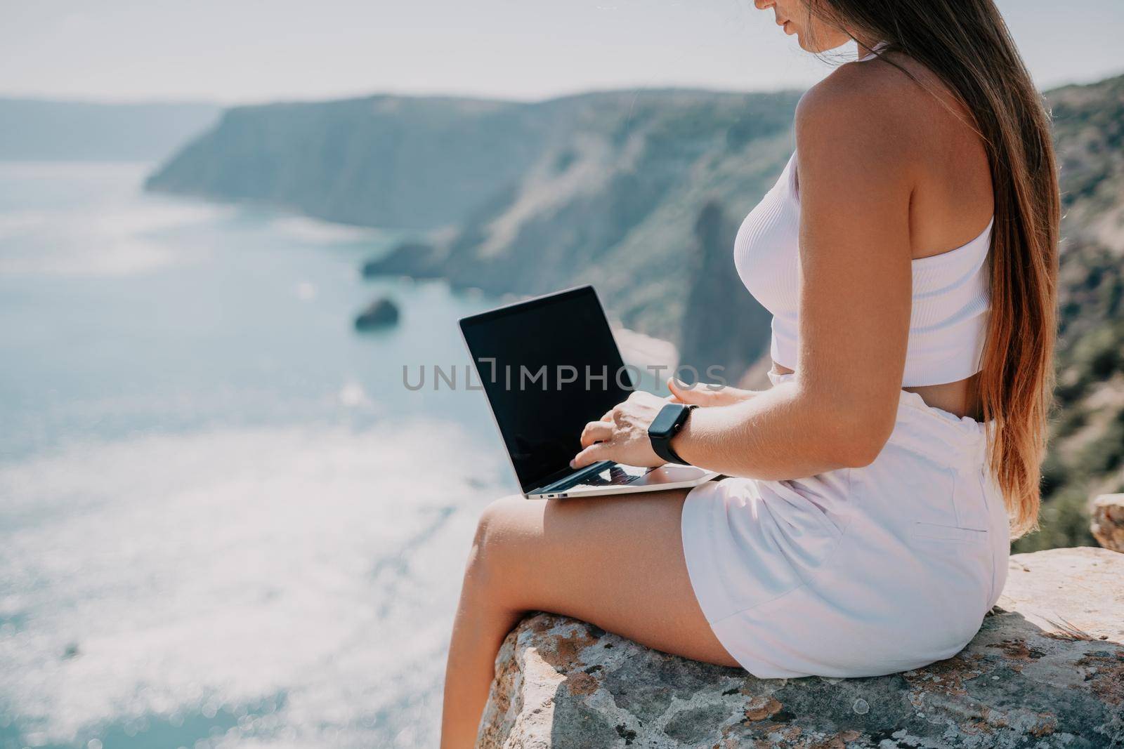 Digital nomad, woman in the hat, a business woman with a laptop sits on the rocks by the sea during sunset, makes a business transaction online from a distance. Freelance, remote work on vacation. by panophotograph