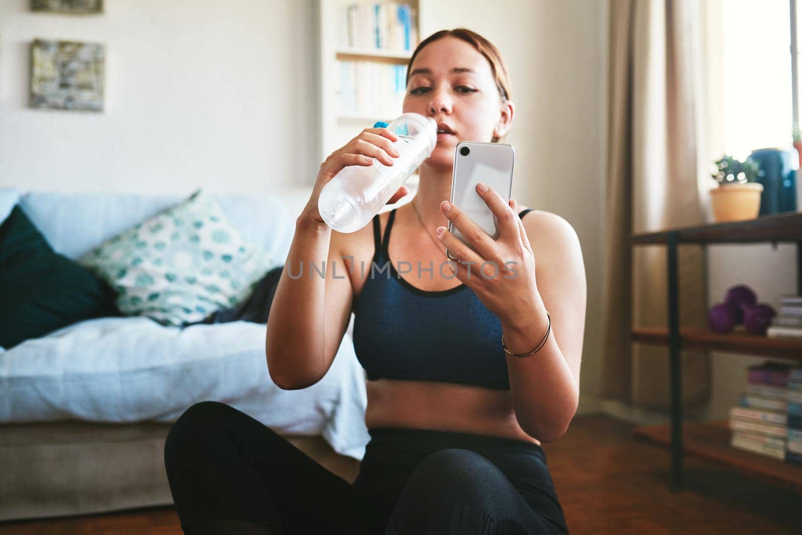 Quick water and social media break. an attractive young woman sitting and drinking water while using her cellphone after a workout. by YuriArcurs