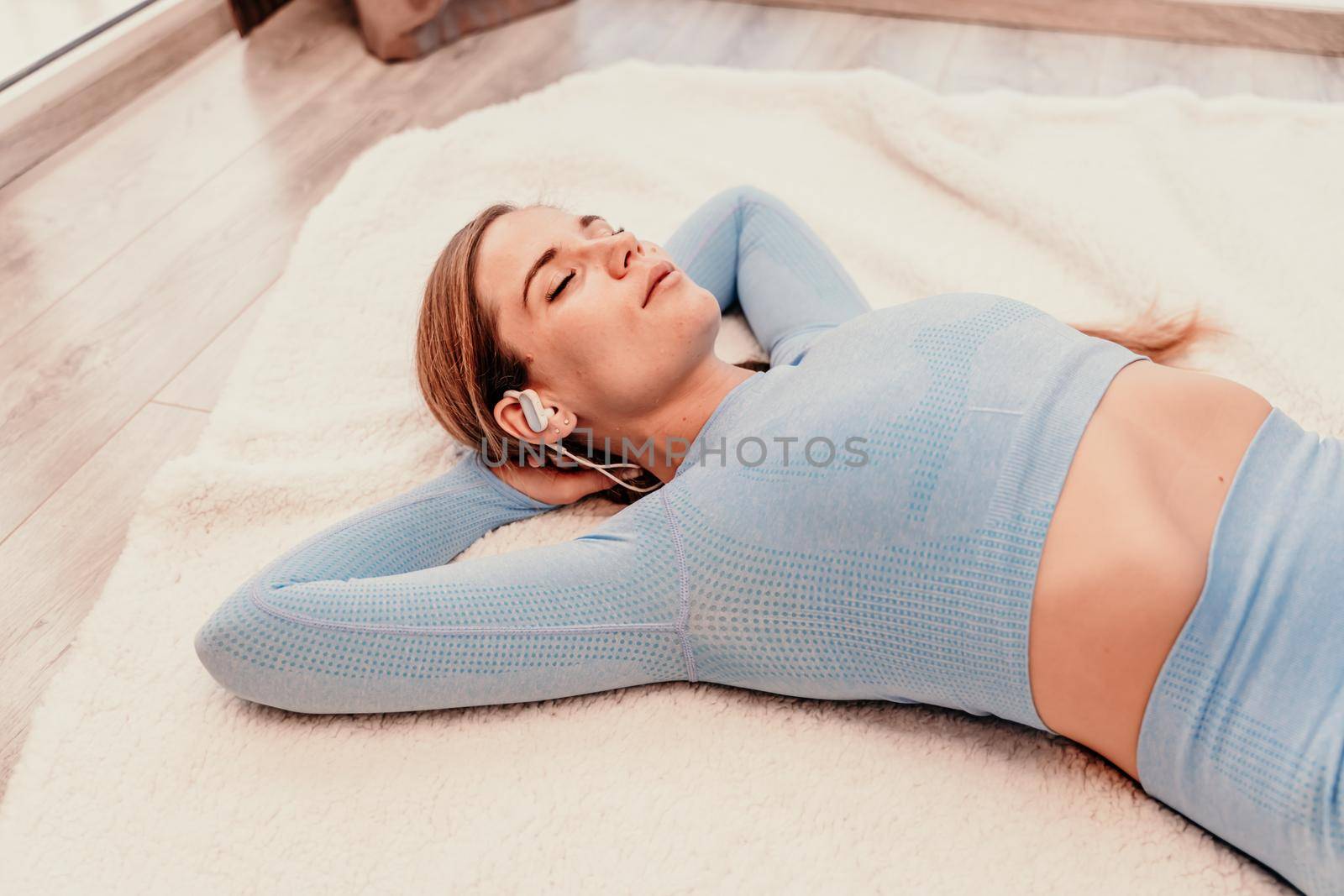 Top view portrait of relaxed woman listening to music with headphones lying on carpet at home. She is dressed in a blue tracksuit