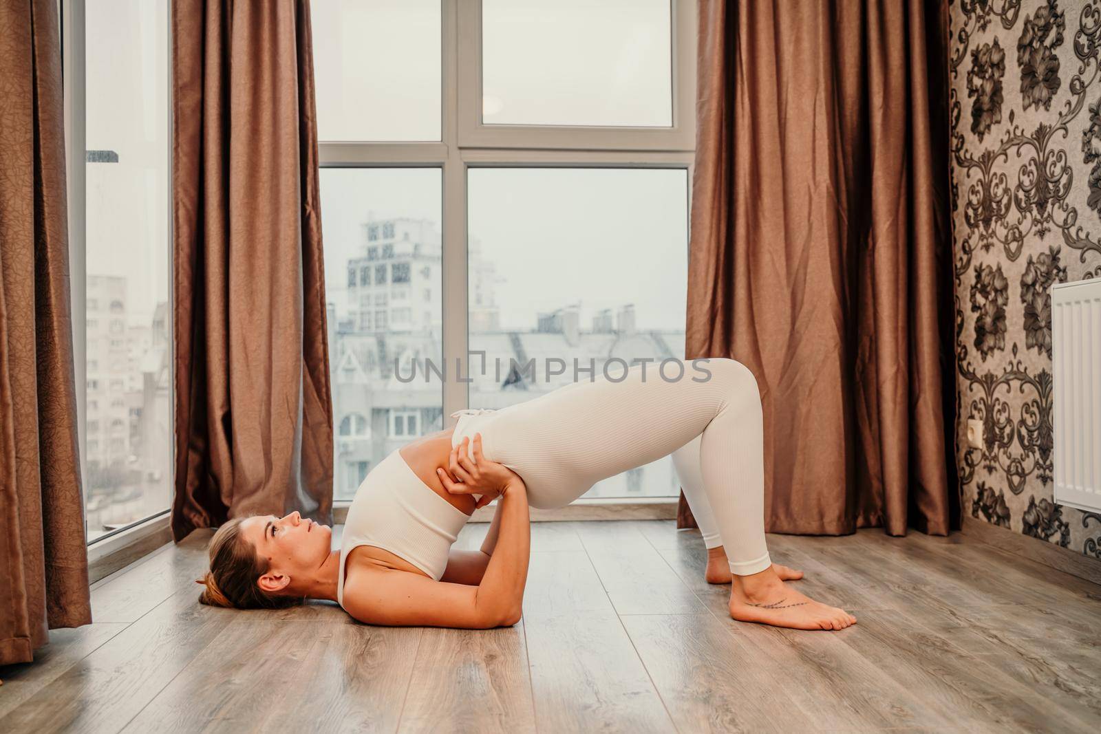 Young athletic attractive woman practicing yoga. Works out at home or in a yoga studio, sportswear, white pants and a full-length top indoors. Healthy lifestyle concept.