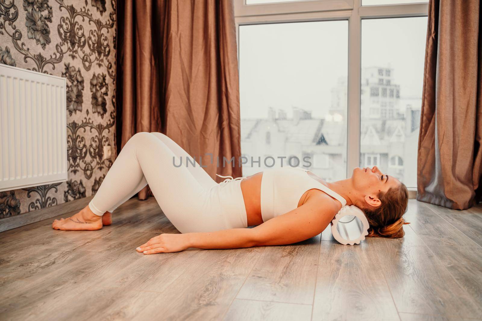 Adult athletic woman, in white bodysuit, performing fascia exercises on the floor - caucasian woman using a massage foam roller - a tool to relieve tension in the back and relieve muscle pain - the concept of physiotherapy and stretching training.