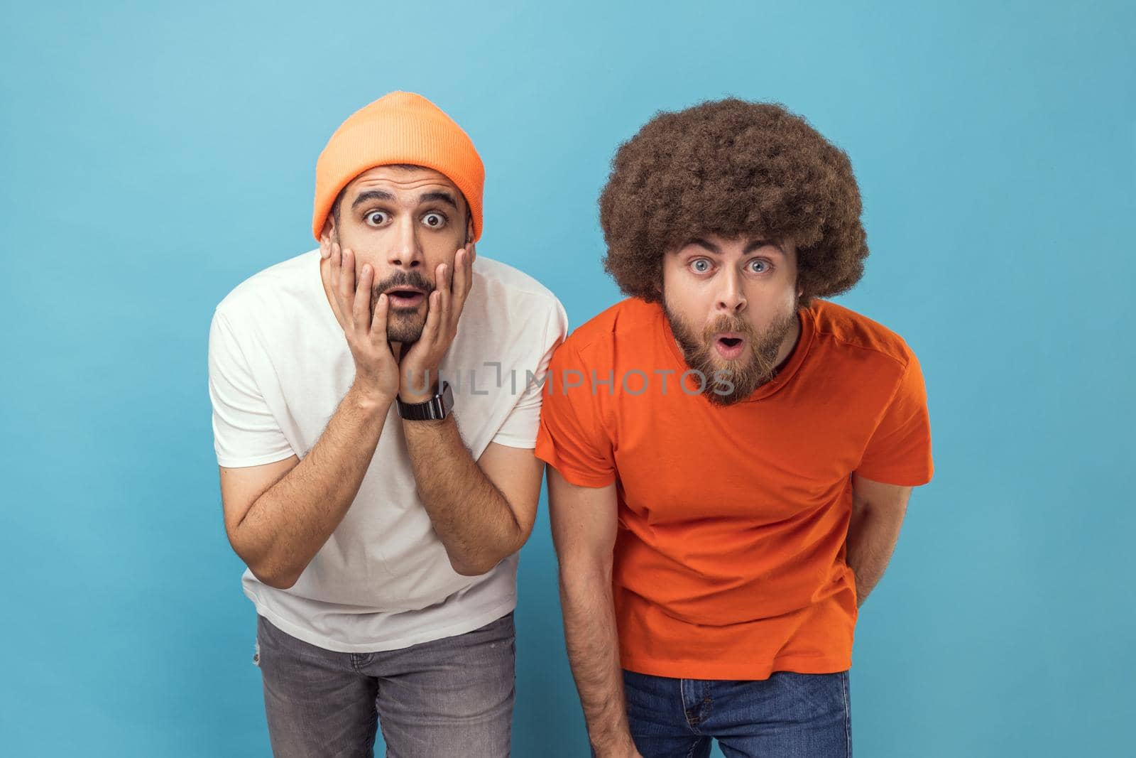 Portrait of two astonished young adult hipster men looking at camera with big eyes and open mouth, shocked by news, sees something amazing. Indoor studio shot isolated on blue background.