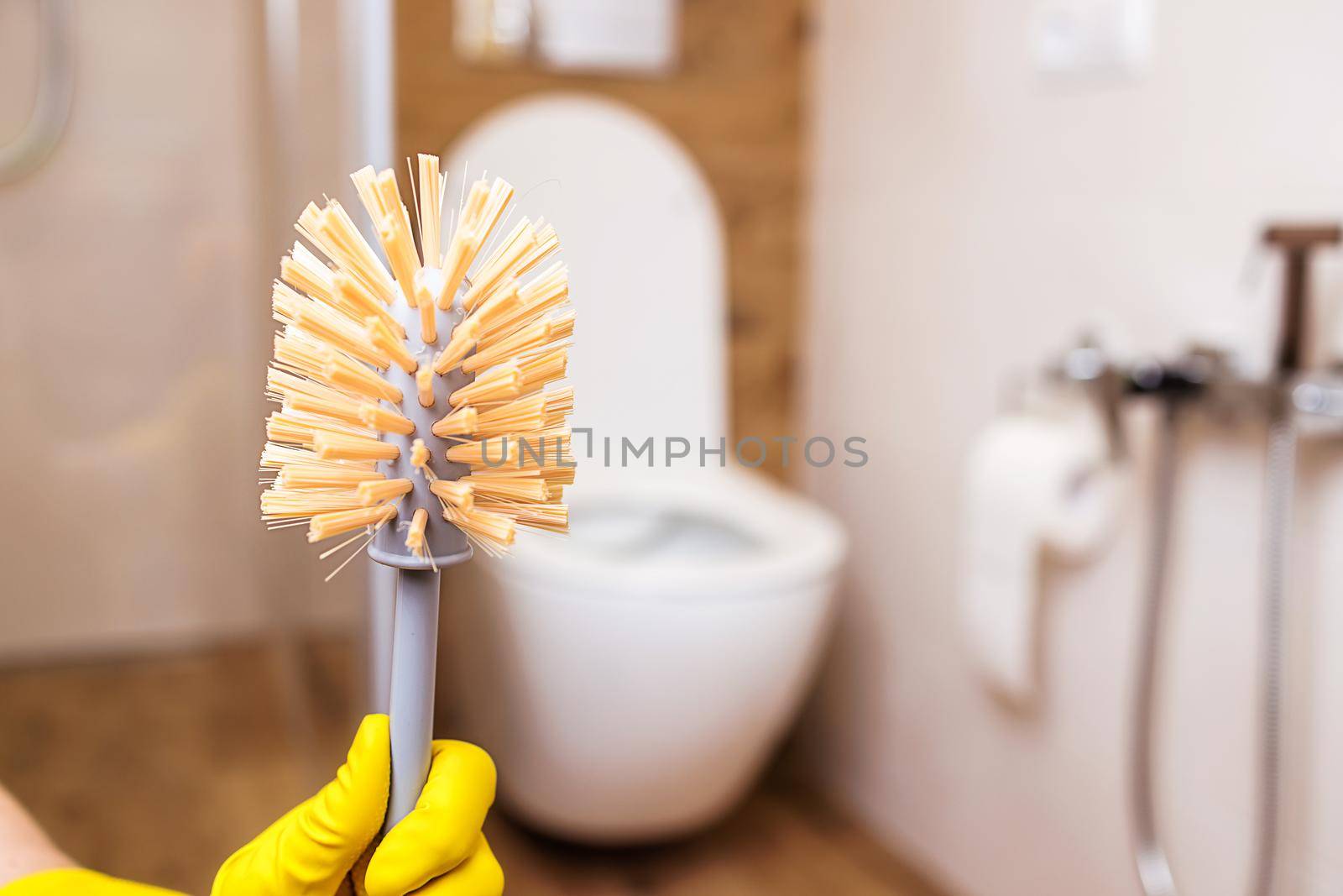 Close-up of a female hand in a yellow rubber cleaning glove holding a clean toilet brush. Beautiful background of a modern bathroom with a white toilet bowl. The concept of hygiene, cleanliness, order