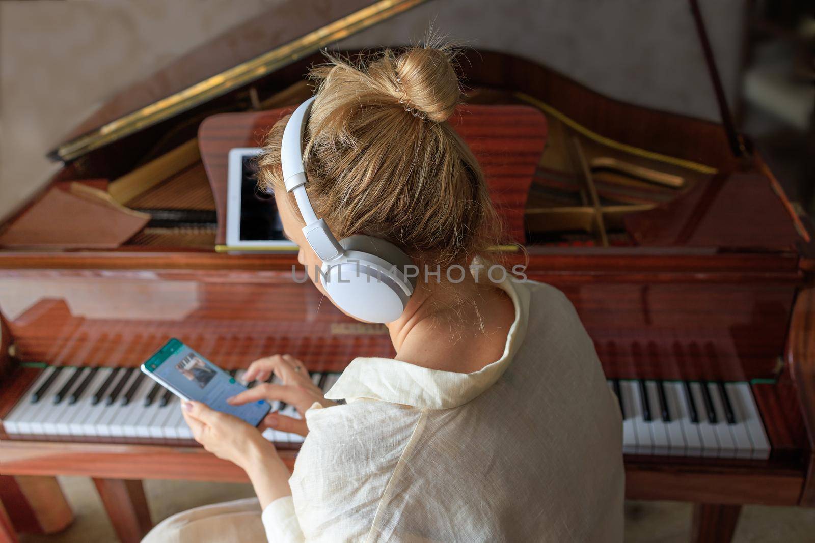 young caucasian blonde girl in headphones plays the vintage piano at home. A woman watches a music lesson on her smartphone. Concept of online learning and new hobby