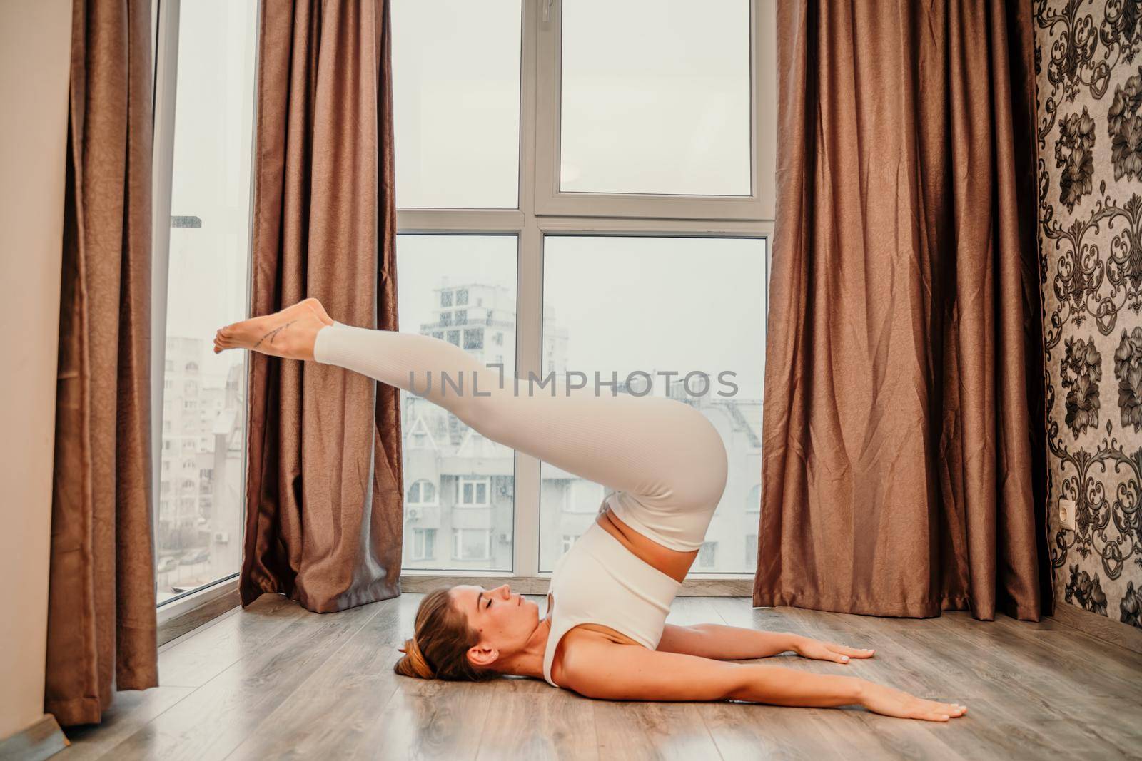 Young athletic attractive woman practicing yoga. Works out at home or in a yoga studio, sportswear, white pants and a full-length top indoors. Healthy lifestyle concept.