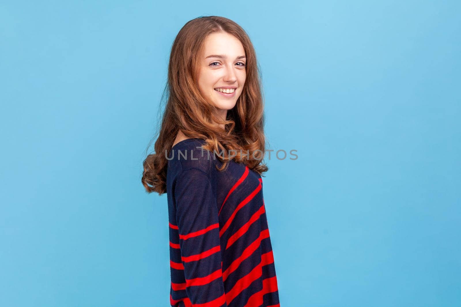 Side view of happy woman wearing striped casual style sweater standing and looking at camera with toothy smile, satisfied expression. Indoor studio shot isolated on blue background.