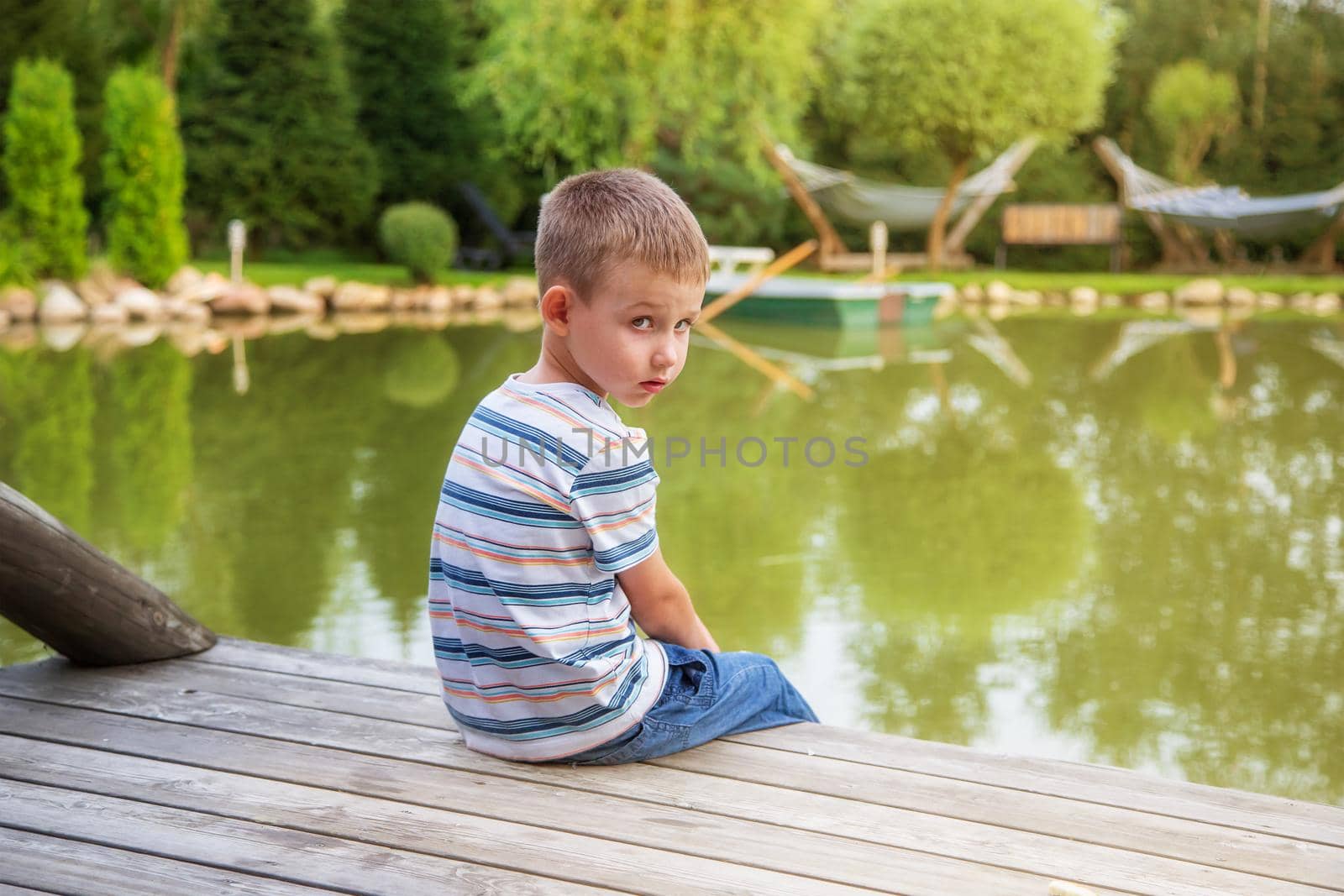 Little European boy in a striped T-shirt, sitting with his back. The child is sad alone. Sunny summer day. The concept of childhood, happiness and tranquility. June 1st,